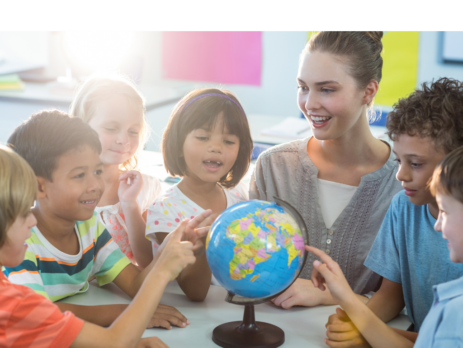 A smiling teacher surrounded by young students smiling and pointing at a globe.